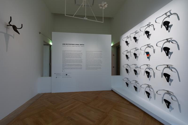 白い展示室の壁に、自転車のサドルとハンドルを組み合わせた展示が並ぶ様子